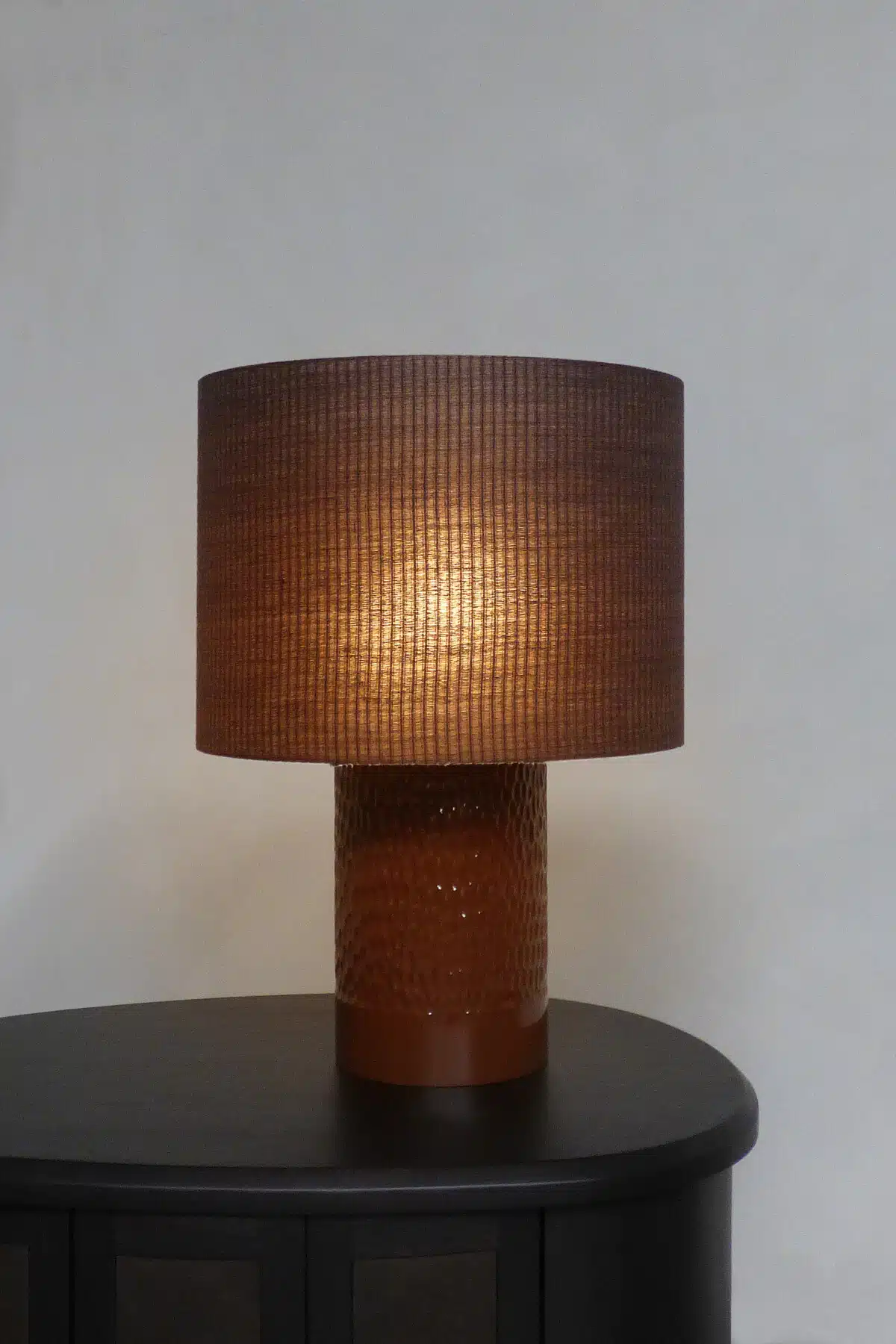 HUGO No. 1 – Handcrafted Belgian table lamp with textured ceramic base and woven drum shade, glowing warmly on a dark side ta