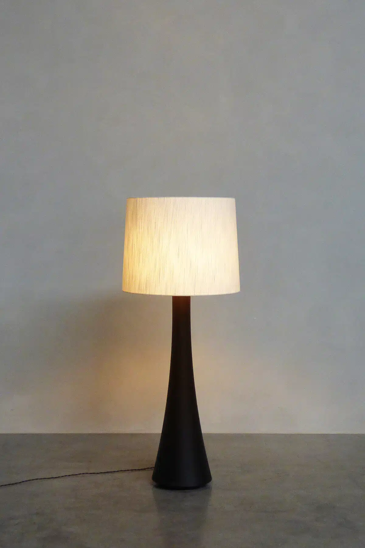 OSCAR NO. 2 – Belgian crafted floor lamp with dark hourglass base and cream textured shade, glowing warmly against a grey wal